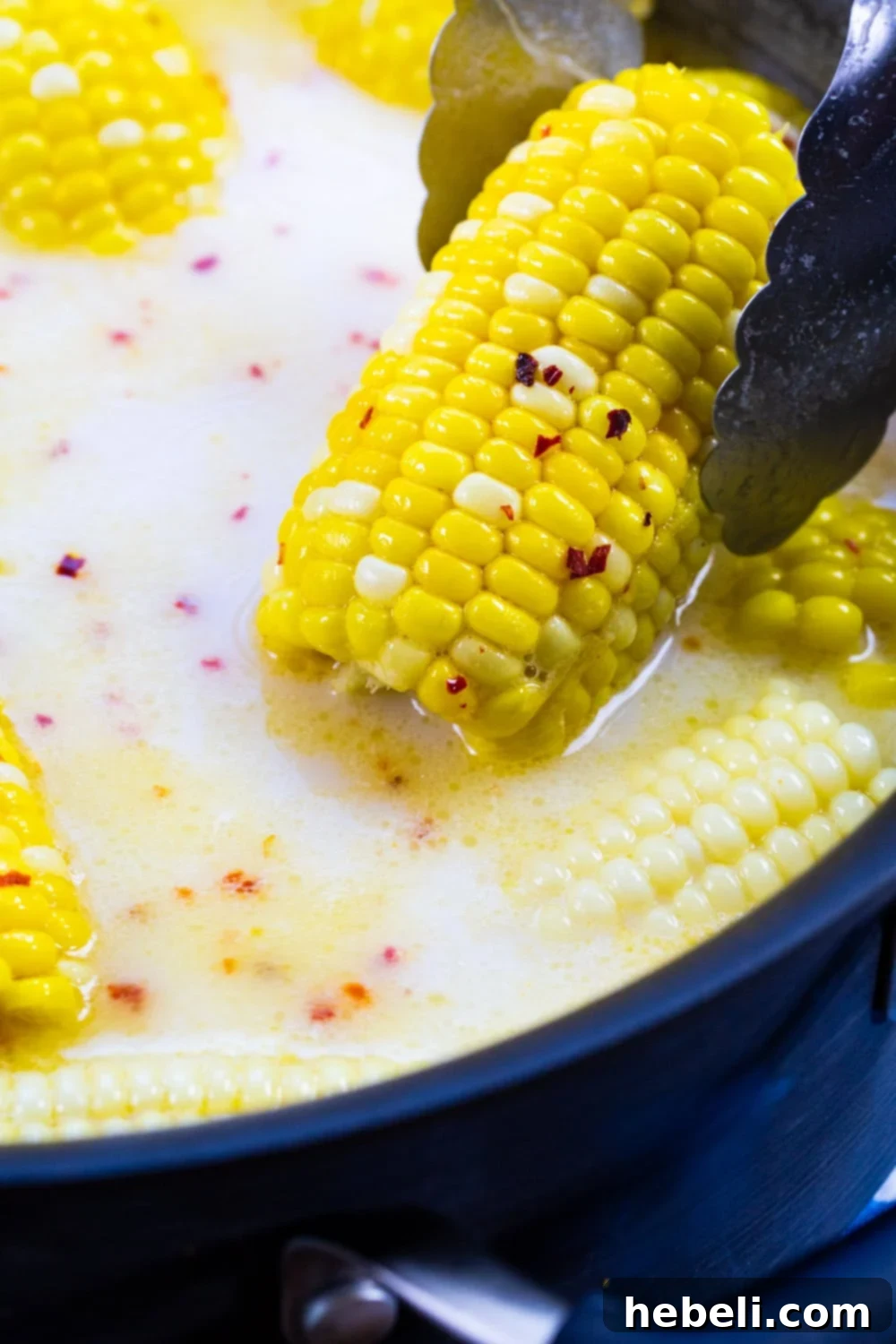 Tongs lifting a piece of perfectly cooked Southern Hot Honey Butter Corn out of the flavorful liquid