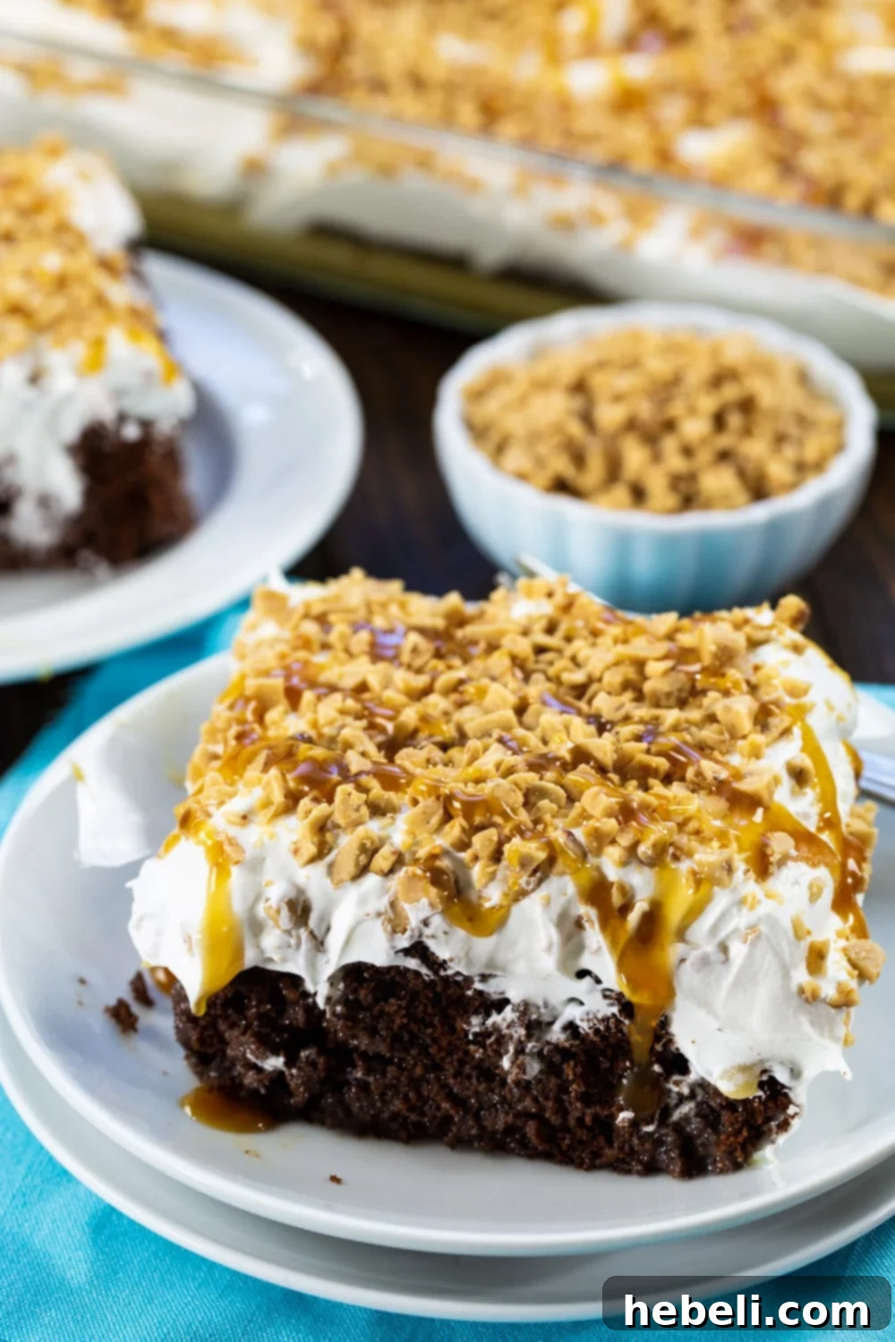 A slice of Toffee Poke Cake with a bowl full of golden toffee bits in the background, ready for topping.