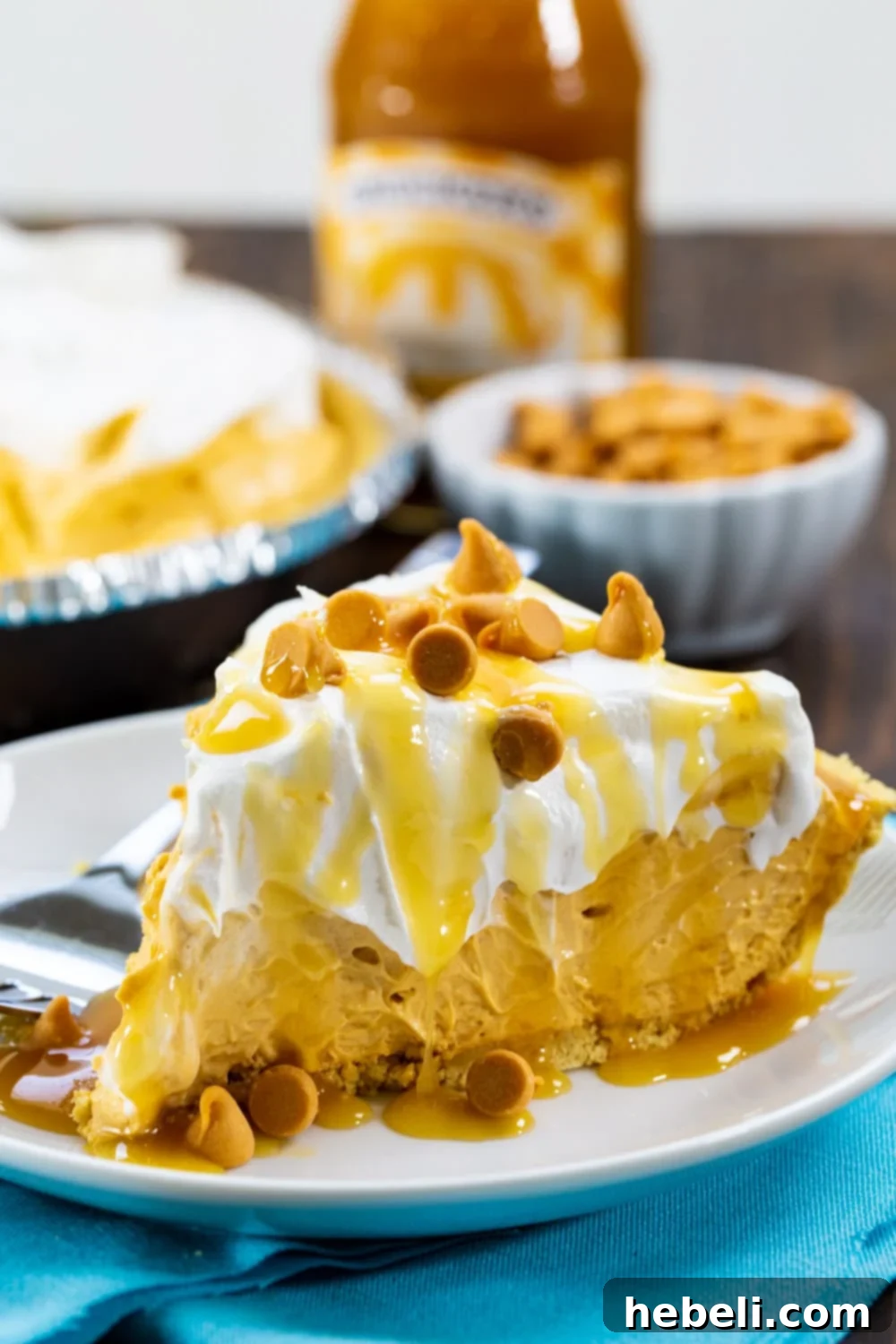 A slice of No-Bake Butterscotch Pie on a plate, with a jar of butterscotch topping and a bowl of butterscotch chips in the soft background.