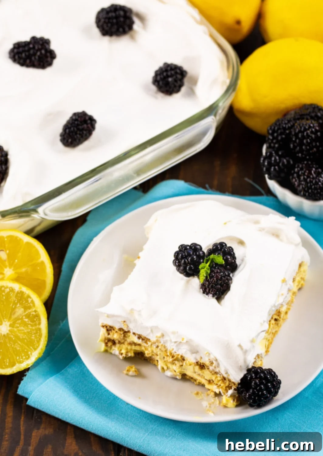 Zesty Lemon Icebox Cake 3 A pristine slice of Lemon Icebox Cake on a white plate, surrounded by whole fresh lemons and the remaining cake in a baking dish.