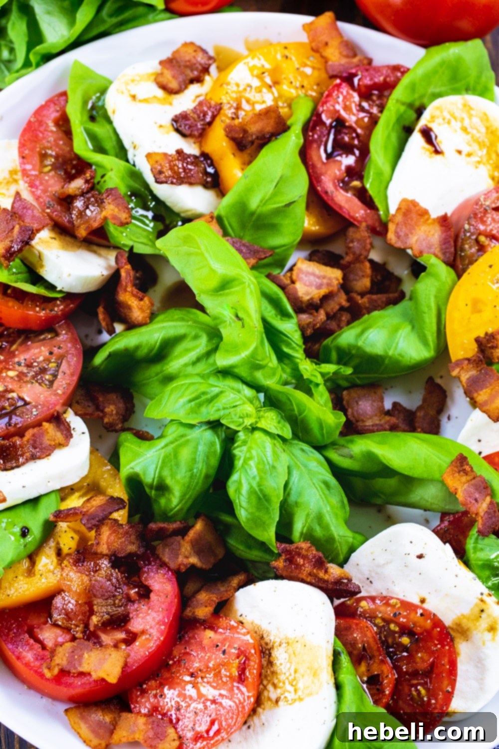 An overhead shot of Bacon Caprese Salad, showcasing the vibrant colors and textures of fresh tomatoes, mozzarella, basil, and crumbled bacon on a large serving plate, with balsamic glaze glistening.