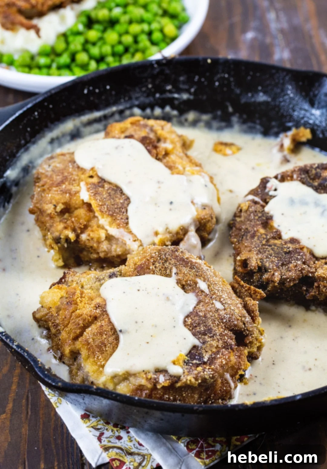 Two Southern Fried Pork Chops with creamy white gravy, fresh herbs, in a rustic cast iron pan.