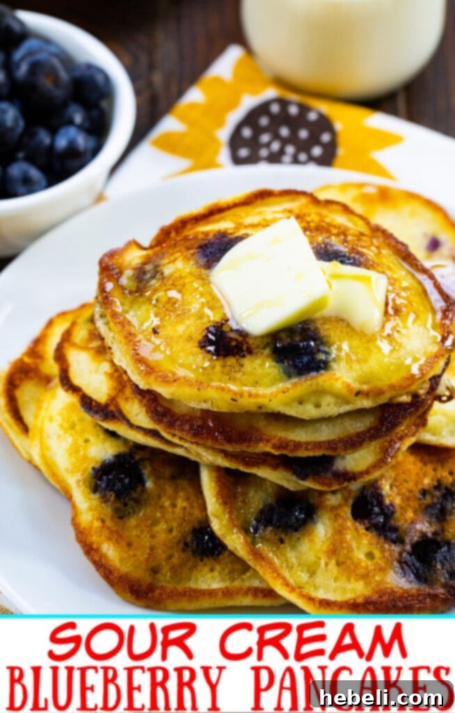 A close-up shot of a perfectly stacked pile of Sour Cream Blueberry Pancakes, glistening with maple syrup and adorned with fresh blueberries.