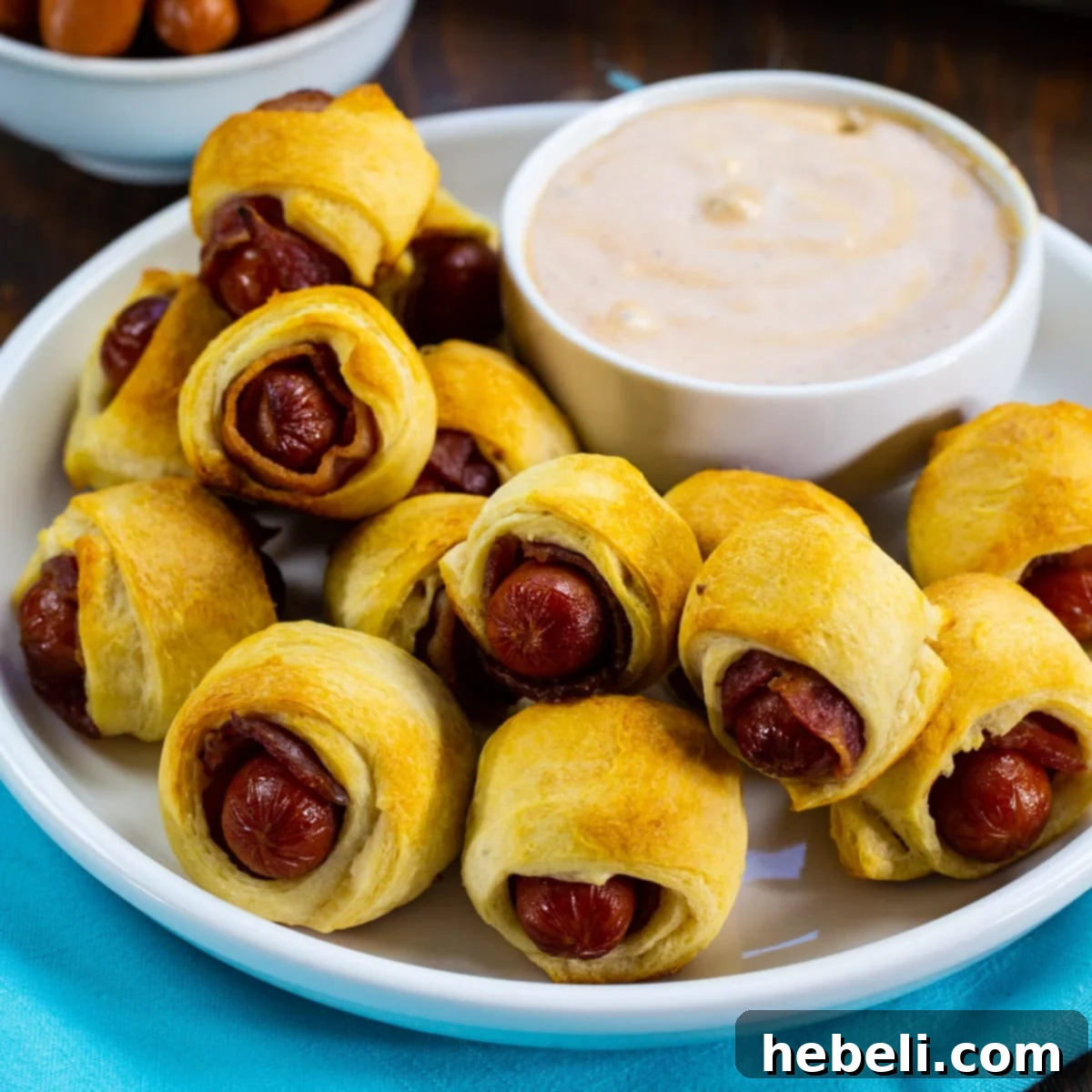 Bacon-Wrapped Sausage Bites 2 Crispy Bacon Pigs in a Blanket served on a rustic plate with a side bowl of creamy BBQ Ranch dipping sauce, garnished and ready for a party.