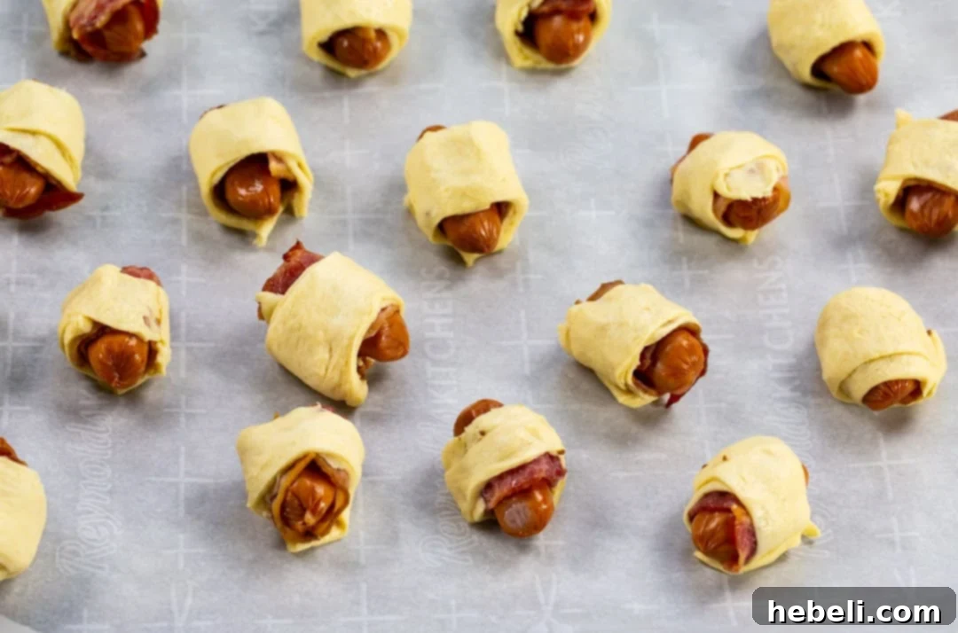 Bacon-Wrapped Sausage Bites 3 Uncooked cocktail wieners, each wrapped in a half-slice of bacon and then snuggly enveloped in strips of crescent dough, arranged on a baking sheet.