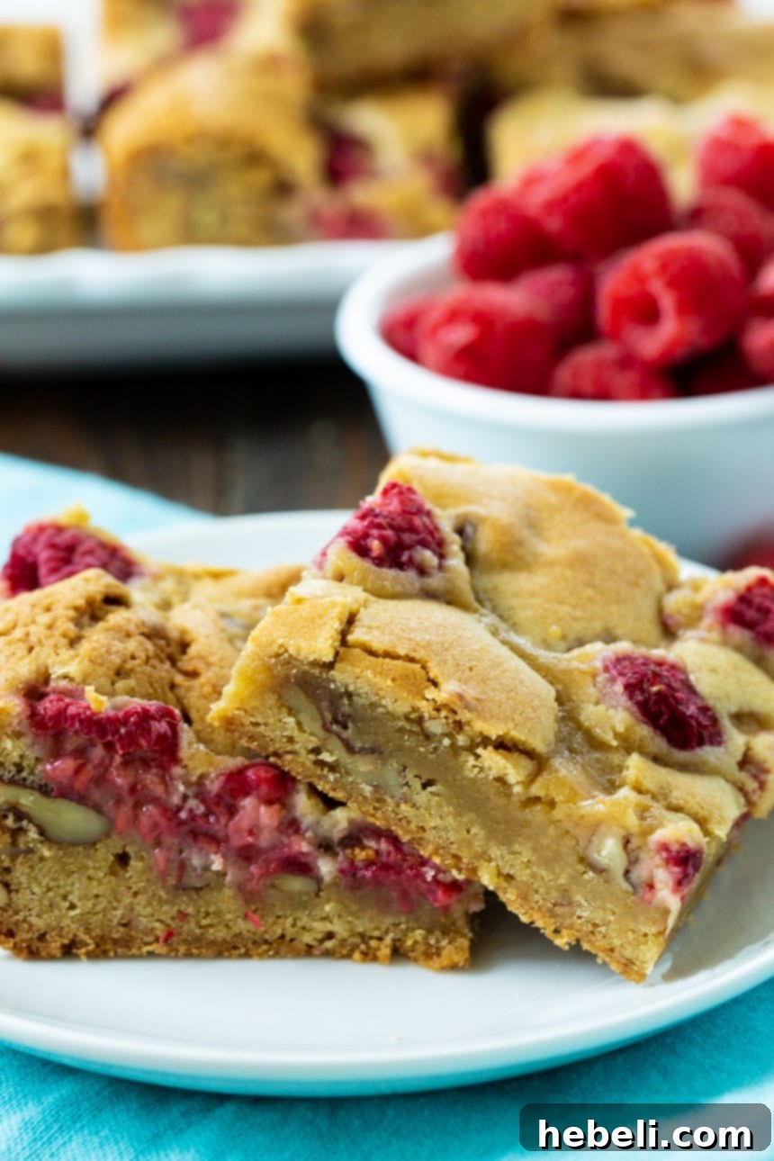 A close-up of Raspberry Pecan Blondies on a plate with more blondies and fresh raspberries in the blurred background, highlighting their deliciousness.