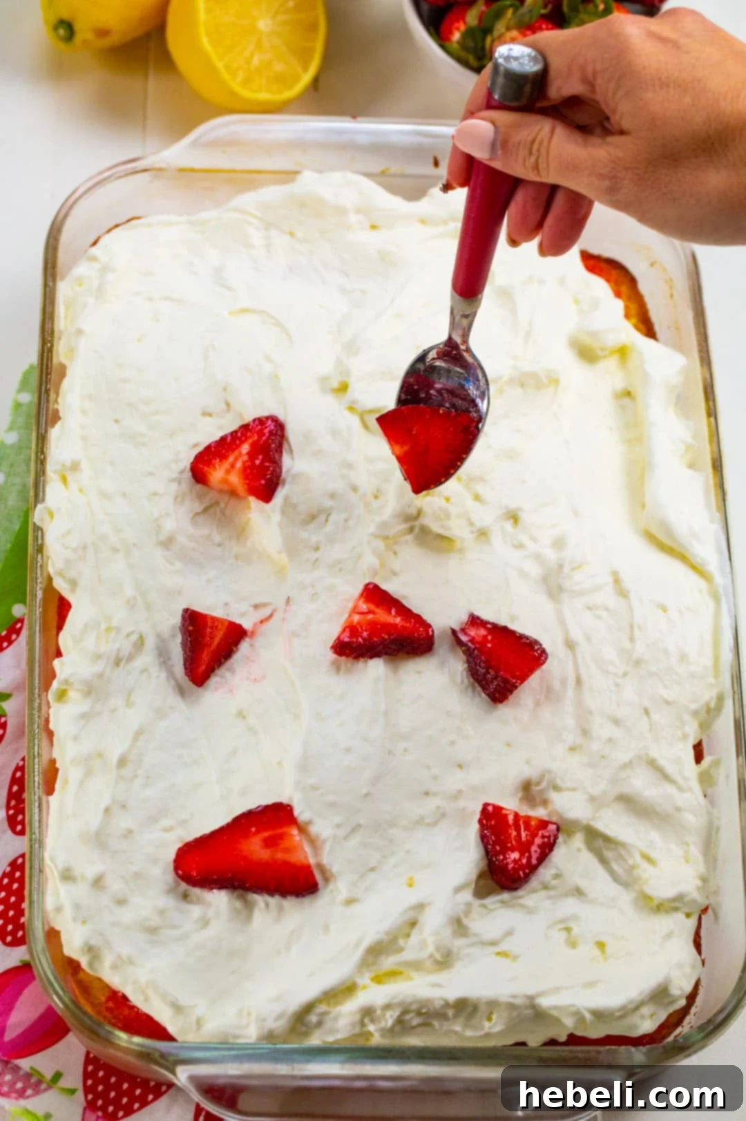 A hand spooning fresh, glistening macerated strawberries over the creamy frosting of the poke cake, adding the final decorative and flavorful touch.