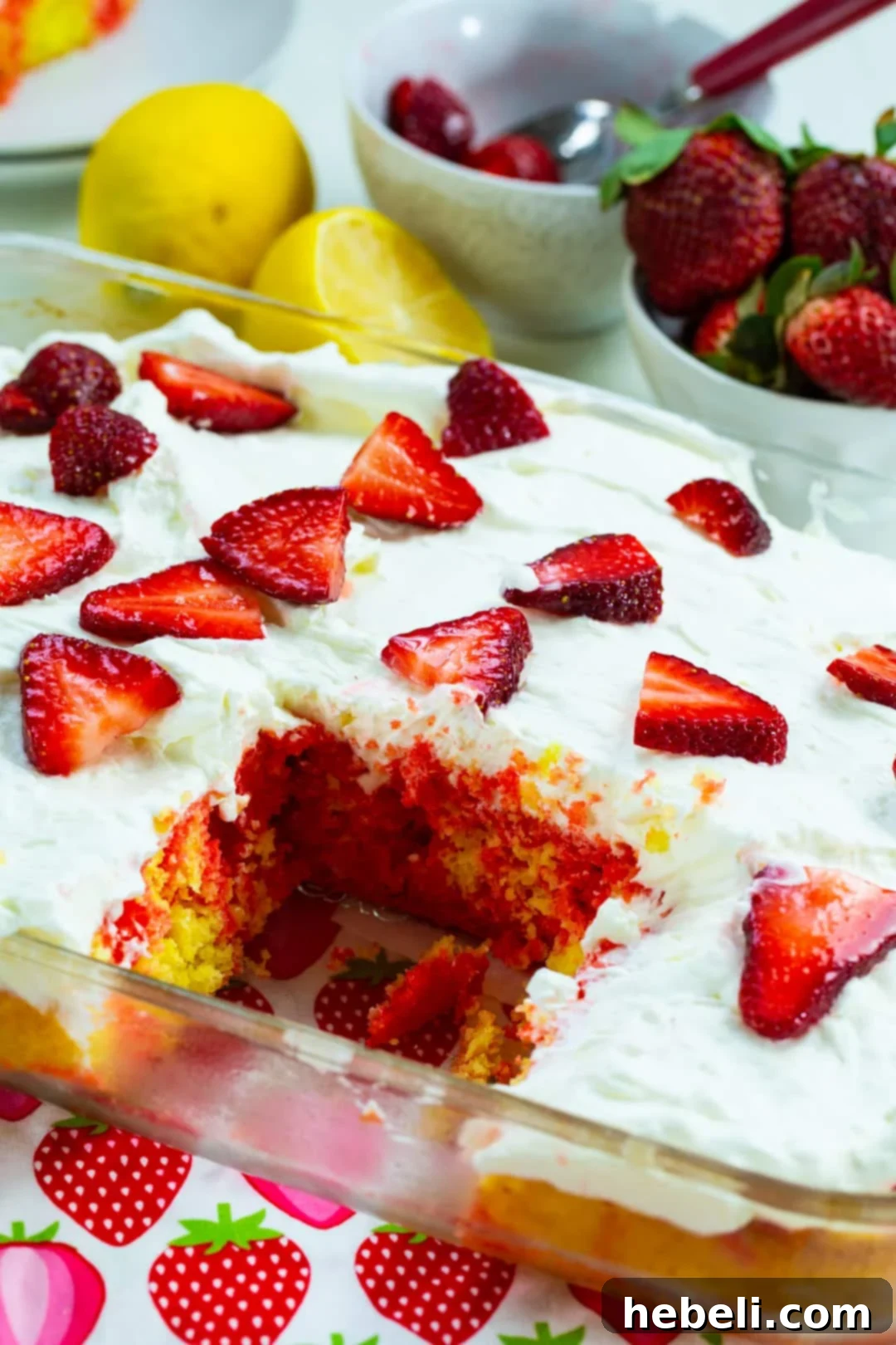 A full Strawberry Lemonade Poke Cake in a baking dish with one slice removed, showcasing the beautiful layers of cake, gelatin, and frosting, garnished with fresh strawberries.