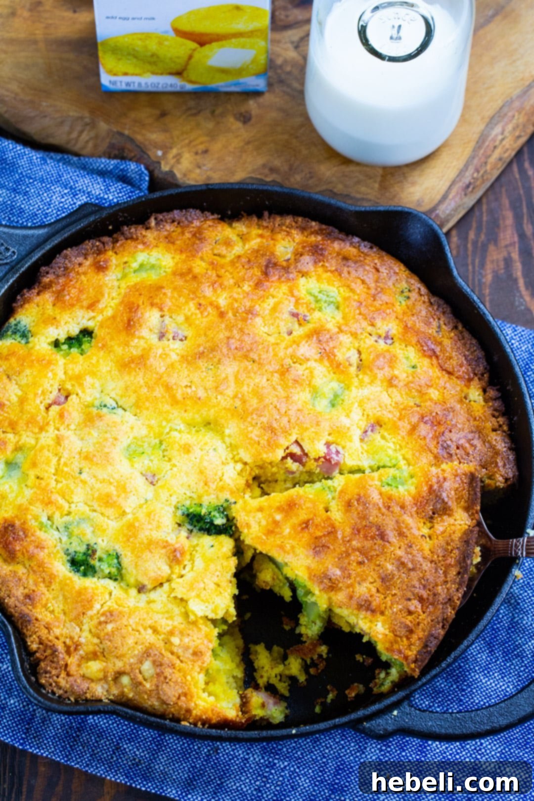A golden-crusted cornbread, featuring visible pieces of broccoli and ham, with a slice removed from the cast iron skillet.