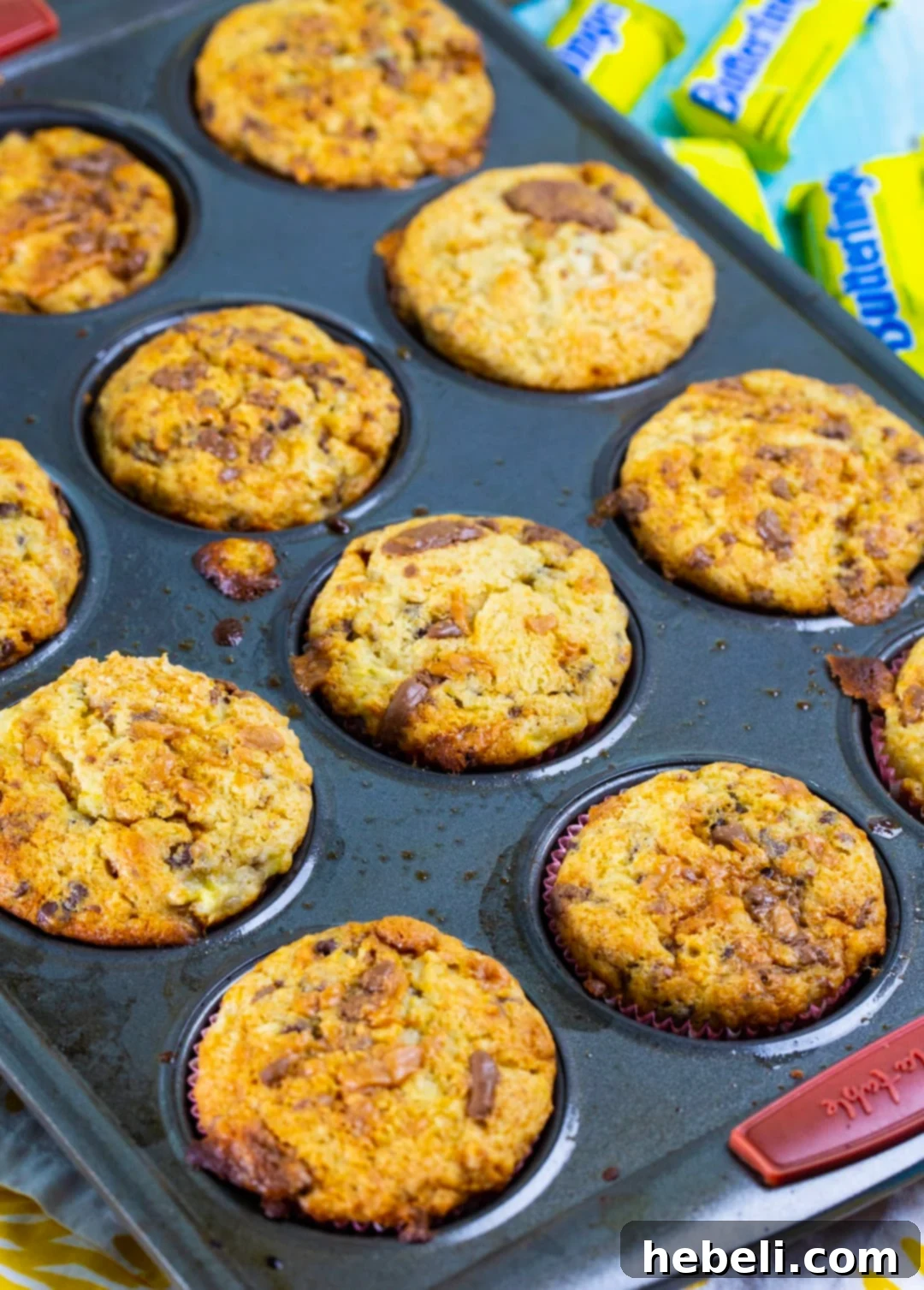 Freshly baked Butterfinger Banana Muffins cooling in a 12-cup muffin tin, their golden domes invitingly showcasing a few visible chocolate chips and candy pieces.
