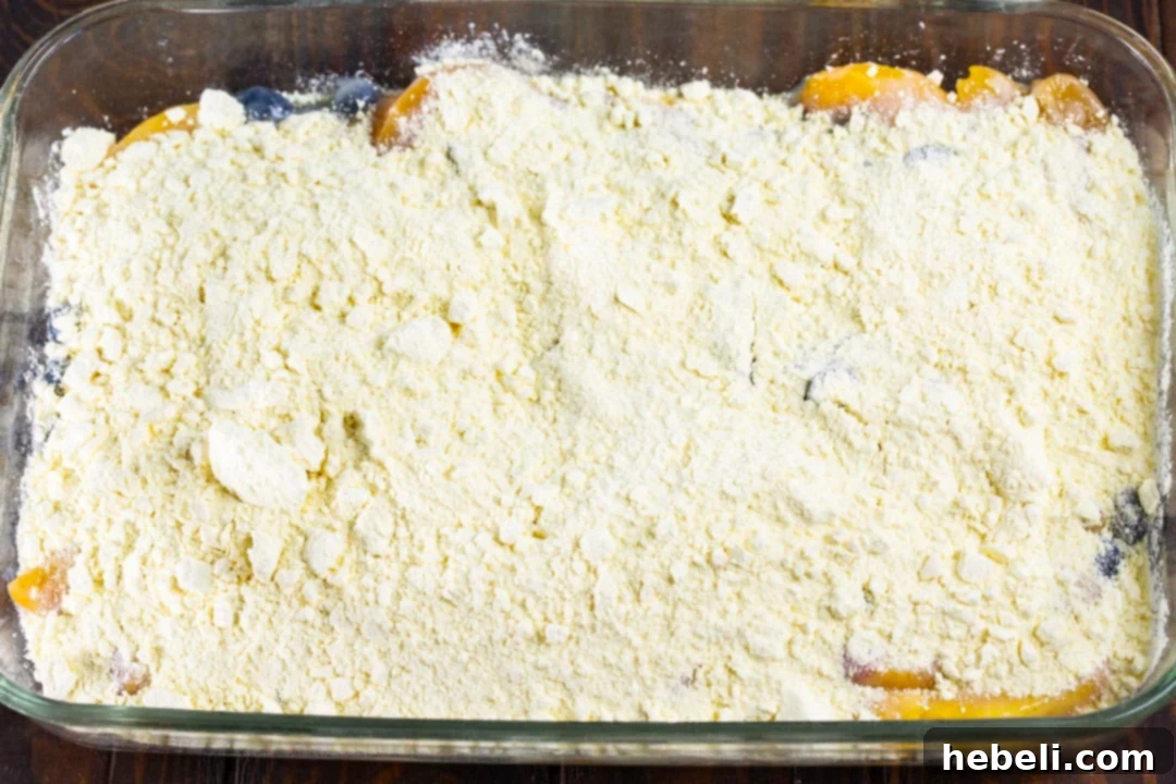 Dry yellow cake mix evenly sprinkled over the sweet blueberry and peach mixture in a baking dish, perfectly poised for the final butter layer to create a golden crust.