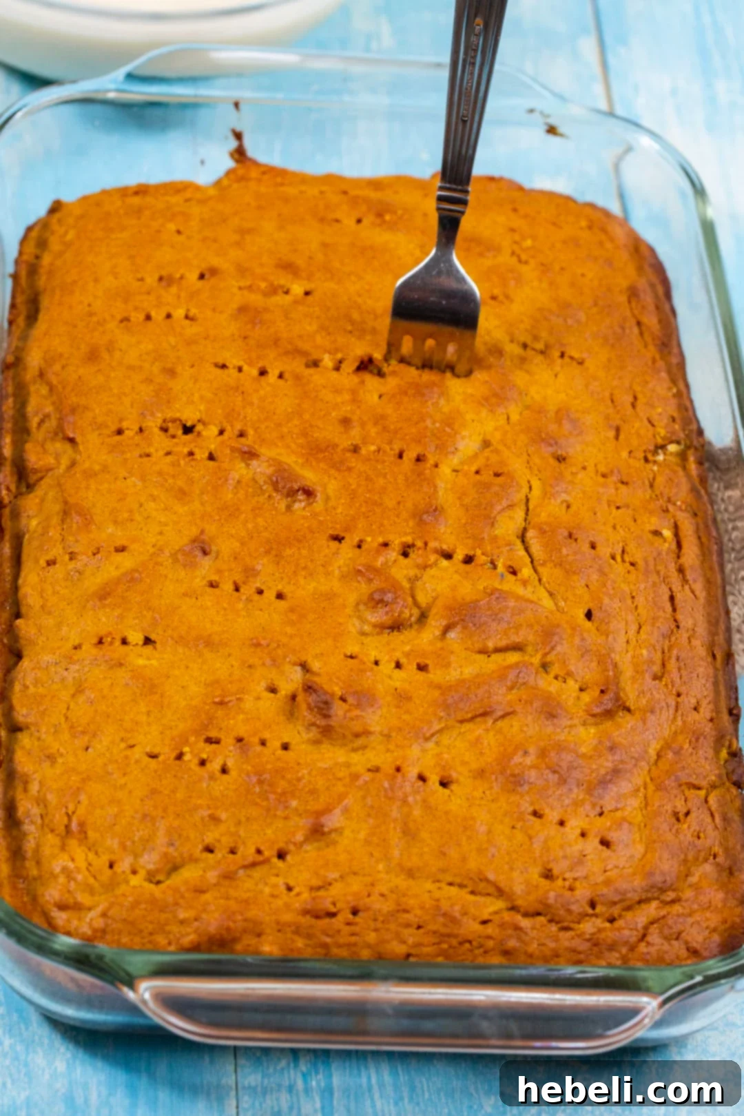 Spiced Pumpkin Tres Leches Cake 3 Close-up shot of a baked cake, with a fork or wooden spoon handle poking holes into its surface, preparing it for the milk soak.