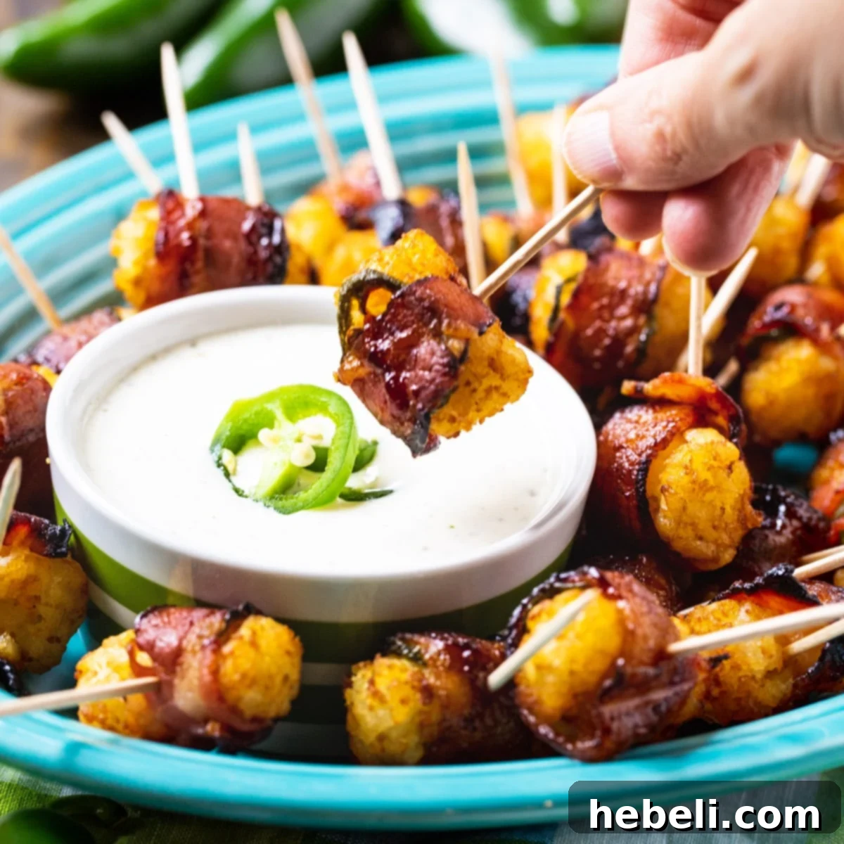 A crispy Jalapeño Bacon Tater Tot being dipped into creamy ranch dressing, highlighting its golden-brown exterior and the vibrant green jalapeño peeking through the bacon wrap.