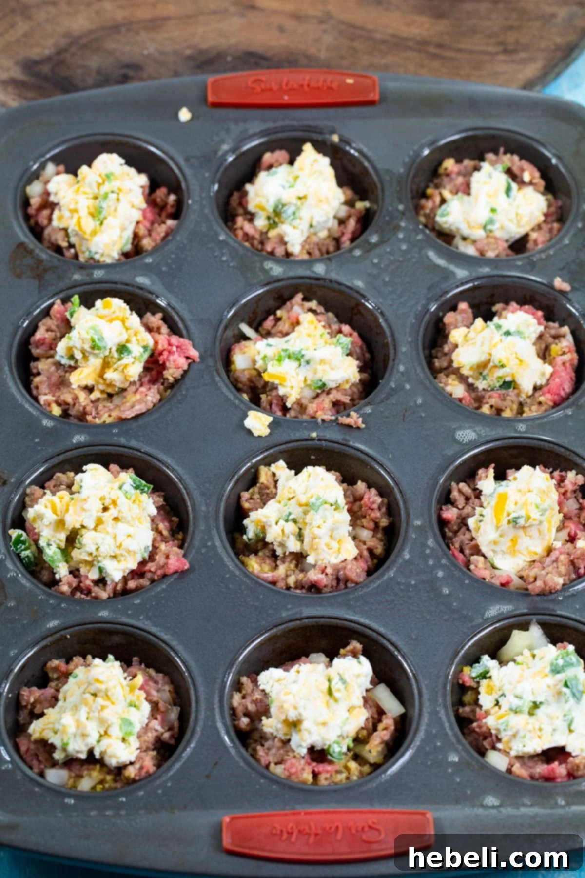 Softened cream cheese mixture being spooned onto the bottom layer of ground beef in a muffin cup.