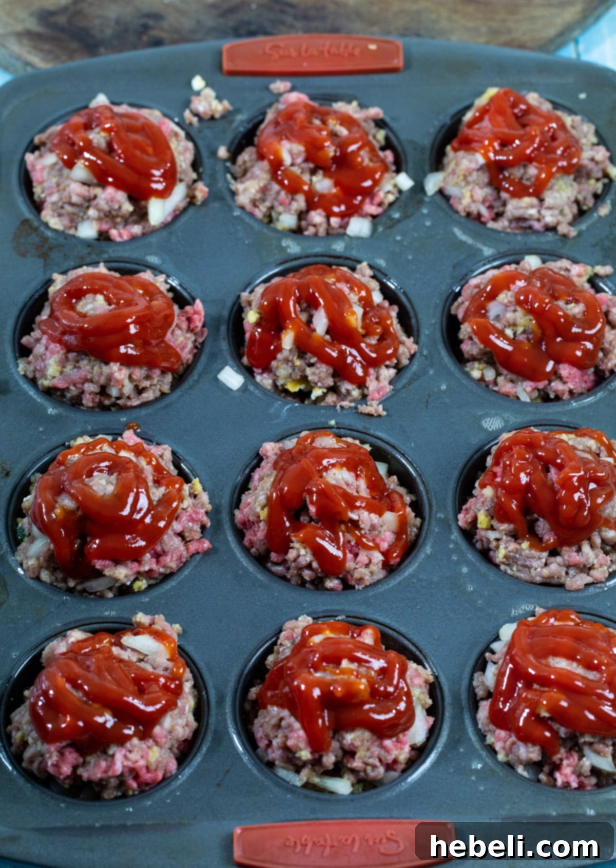 Ketchup being generously squirted over the tops of unbaked meatloaf muffins.