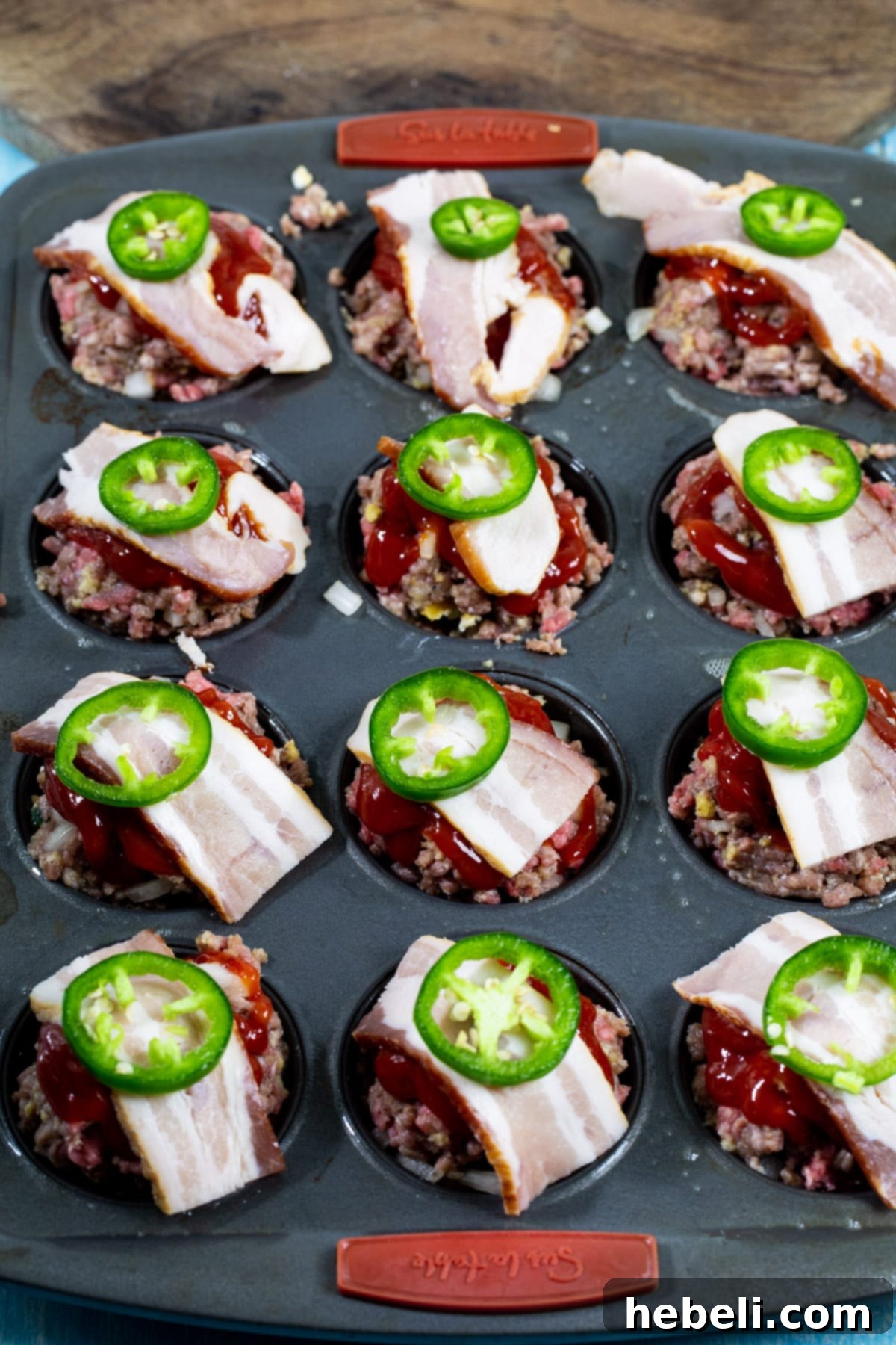 Bacon pieces and a slice of fresh jalapeno placed as toppings on unbaked meatloaf muffins.