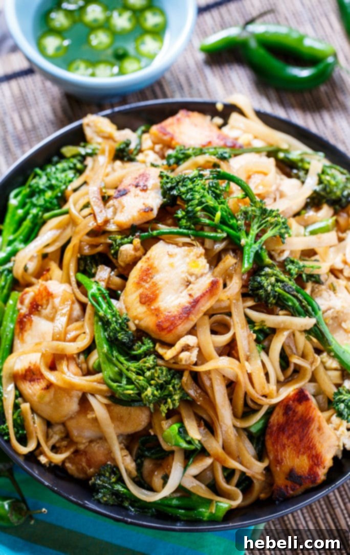 A black bowl brimming with Thai Stir-Fried Noodles, chicken, and vibrant broccolini, garnished with fresh cilantro and served alongside sliced jalapenos for an extra kick, perfectly showcasing the dish.