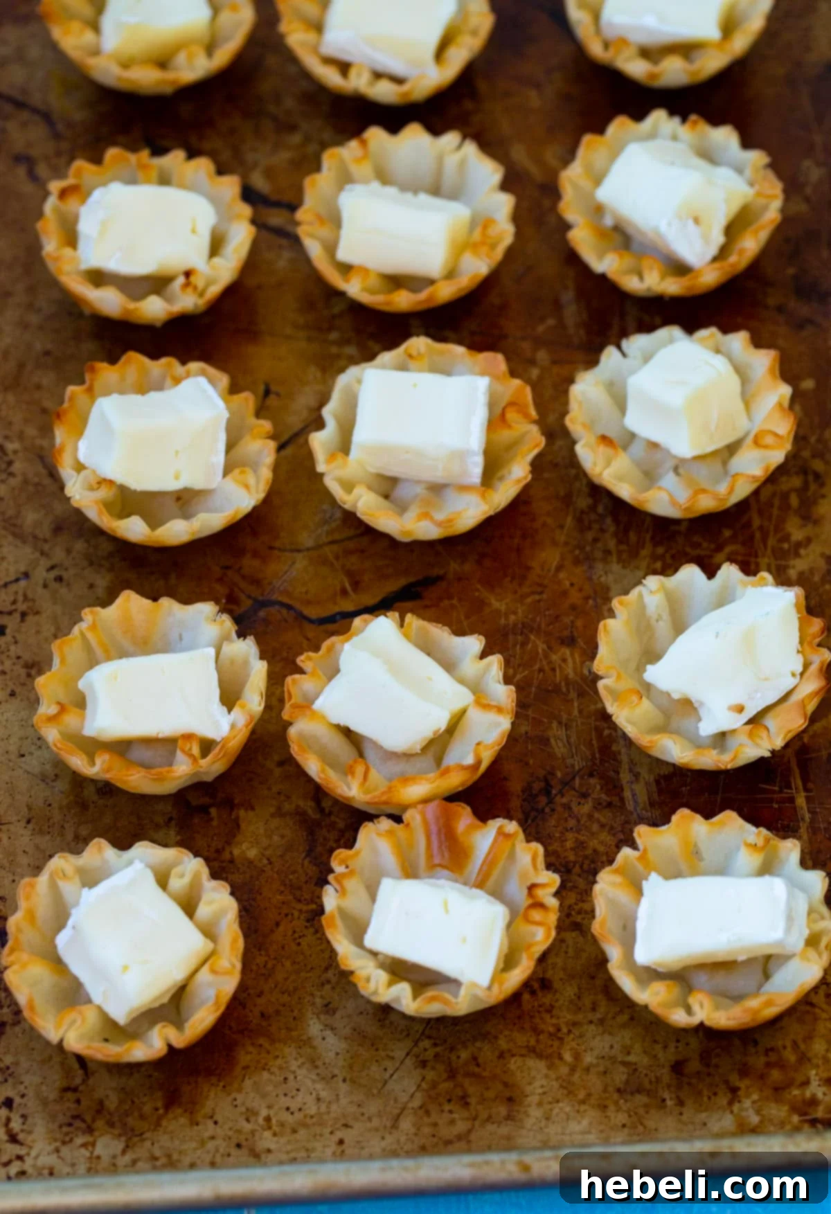 Mini phyllo shells lined up on a baking sheet, each carefully filled with a cube of soft brie cheese, awaiting the next delicious topping.