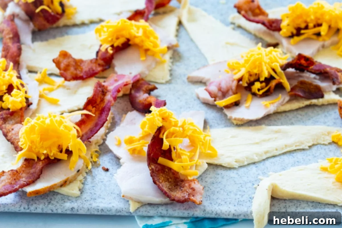 Kentucky Hot Brown Crescent Bake 5 Unrolled crescent dough triangles being topped with slices of turkey, crispy bacon, and a sprinkle of shredded cheddar cheese, ready to be rolled.