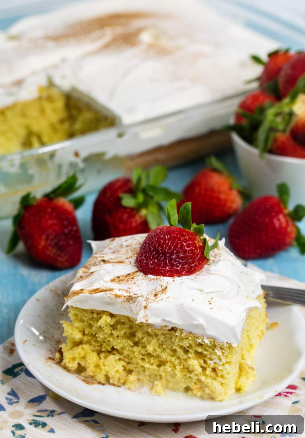 Effortless Tres Leches Cake 3 A beautifully presented slice of Tres Leches cake on a white plate, with the remainder of the cake in a baking dish visible in the soft-focus background.