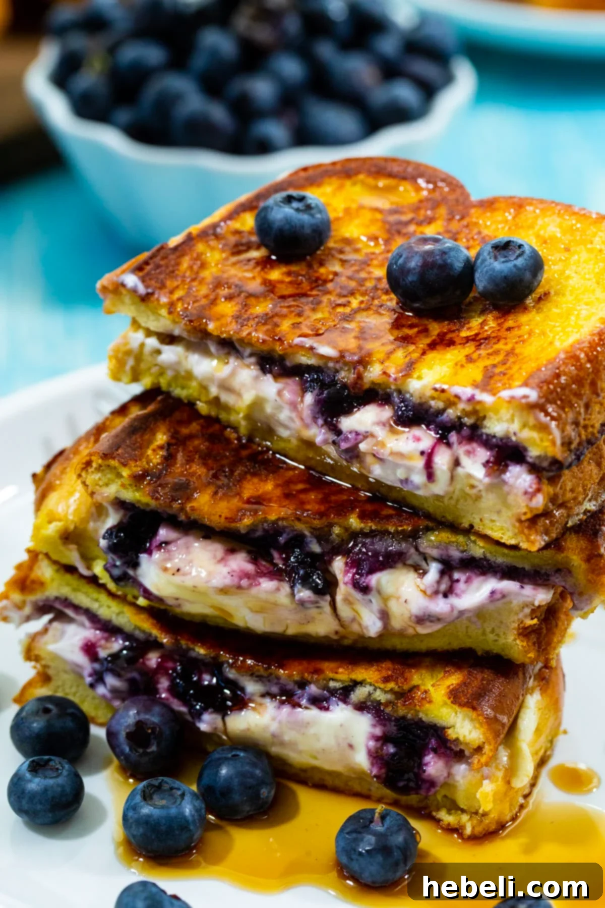 Two halves of Blueberry Stuffed French Toast, perfectly stacked and generously drizzled with glistening maple syrup, adorned with fresh blueberries on a plate.