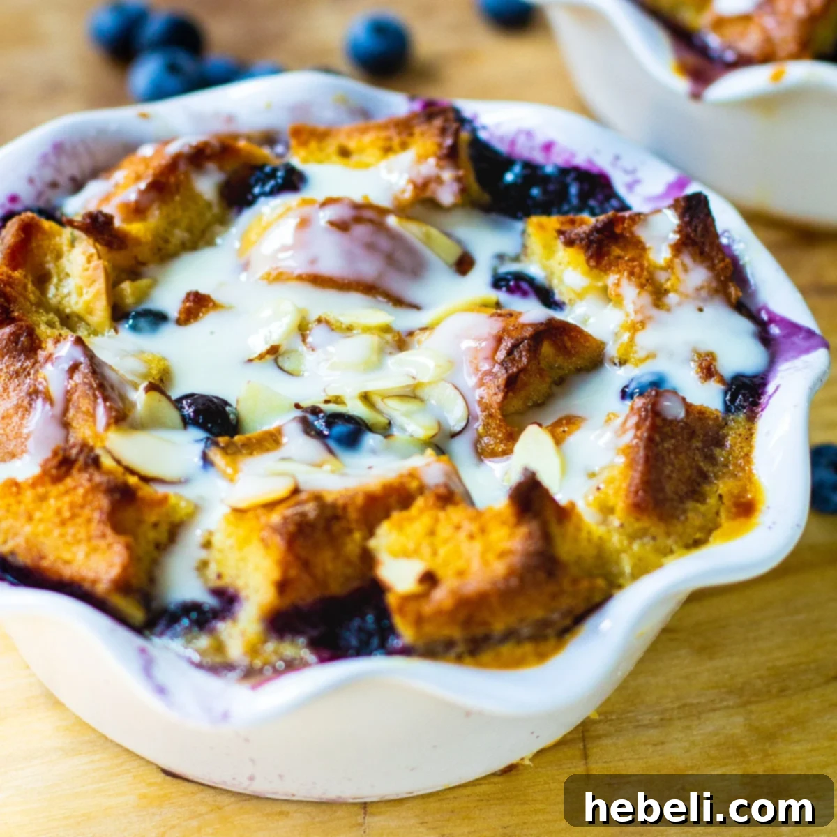 A close-up of individual Blueberry White Chocolate Bread Pudding served in a white ramekin, garnished with fresh blueberries and a generous drizzle of Amaretto Cream Sauce.