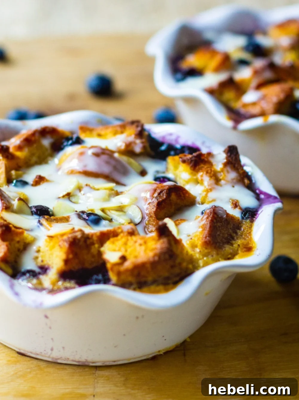 Two white ramekins showcasing individual servings of Blueberry White Chocolate Bread Pudding, adorned with fresh blueberries and a decadent Amaretto Cream Sauce.