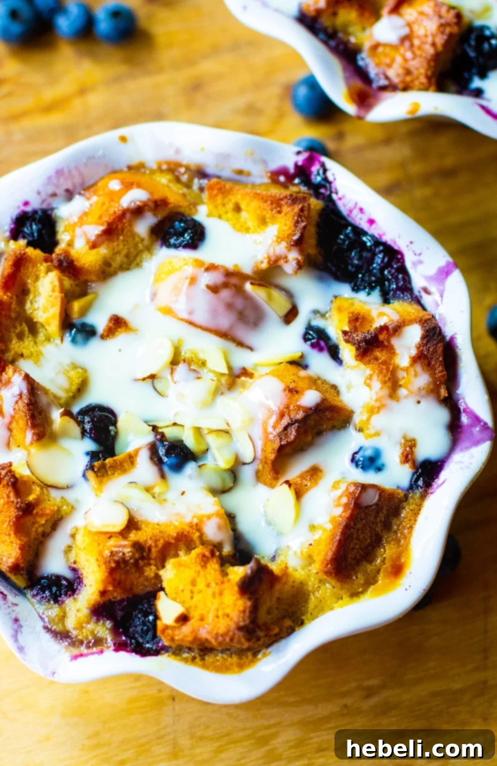 An overhead view of a golden-brown Blueberry White Chocolate Bread Pudding in a baking dish, beautifully drizzled with Amaretto Cream Sauce and scattered with sliced almonds.