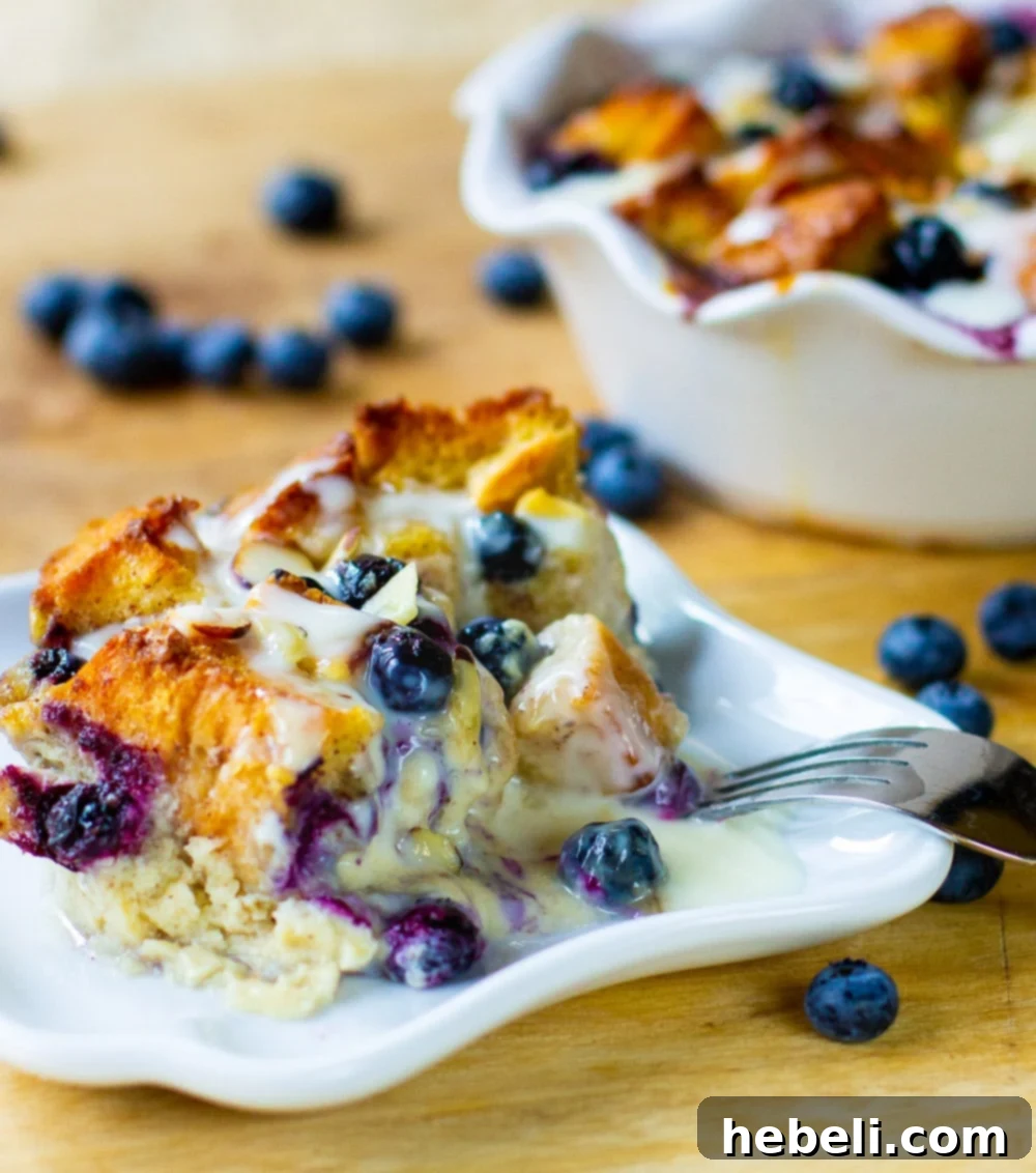 A delectable slice of Blueberry White Chocolate Bread Pudding on a white plate, revealing the layers of bread, blueberries, and melted white chocolate, generously covered in Amaretto Cream Sauce.