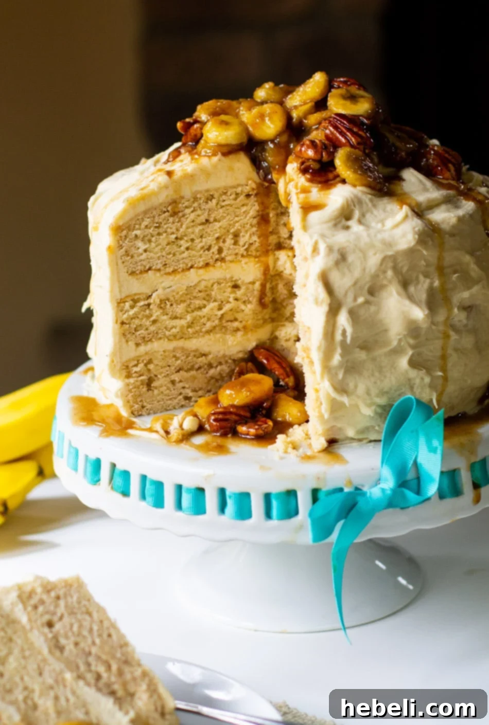 A large, decadent Bananas Foster Cake with caramel cream cheese frosting and banana topping, sitting on a round cake stand.