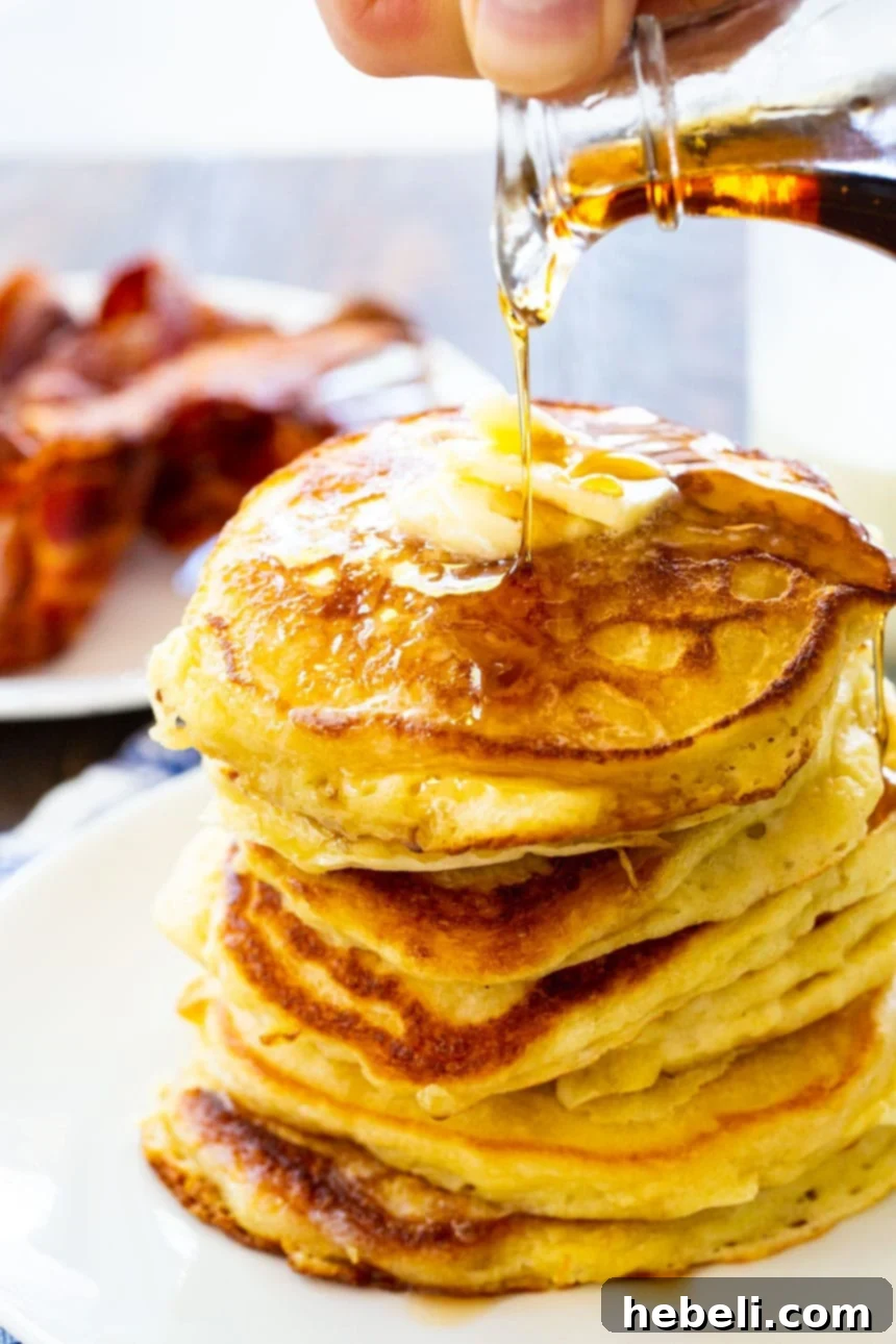 Fluffy Buttermilk Pancakes 8 A tall stack of classic buttermilk pancakes, with crispy bacon blurred in the background, a perfect hearty breakfast.