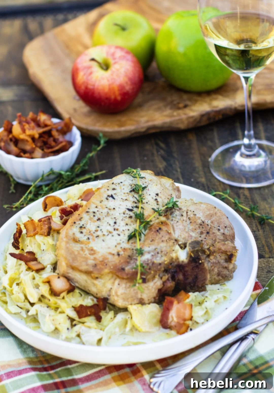 A serving of pork chop over a vibrant bed of creamy cabbage, with sliced apples and a glass of red wine in the background, set on a dark wooden table.