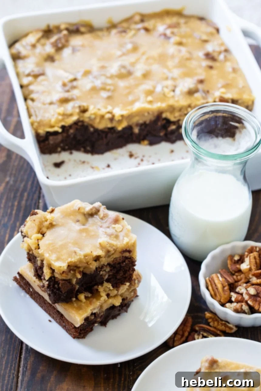 Two Praline Brownies stacked on a plate, with a jug of milk and a pan full of brownies in the background.