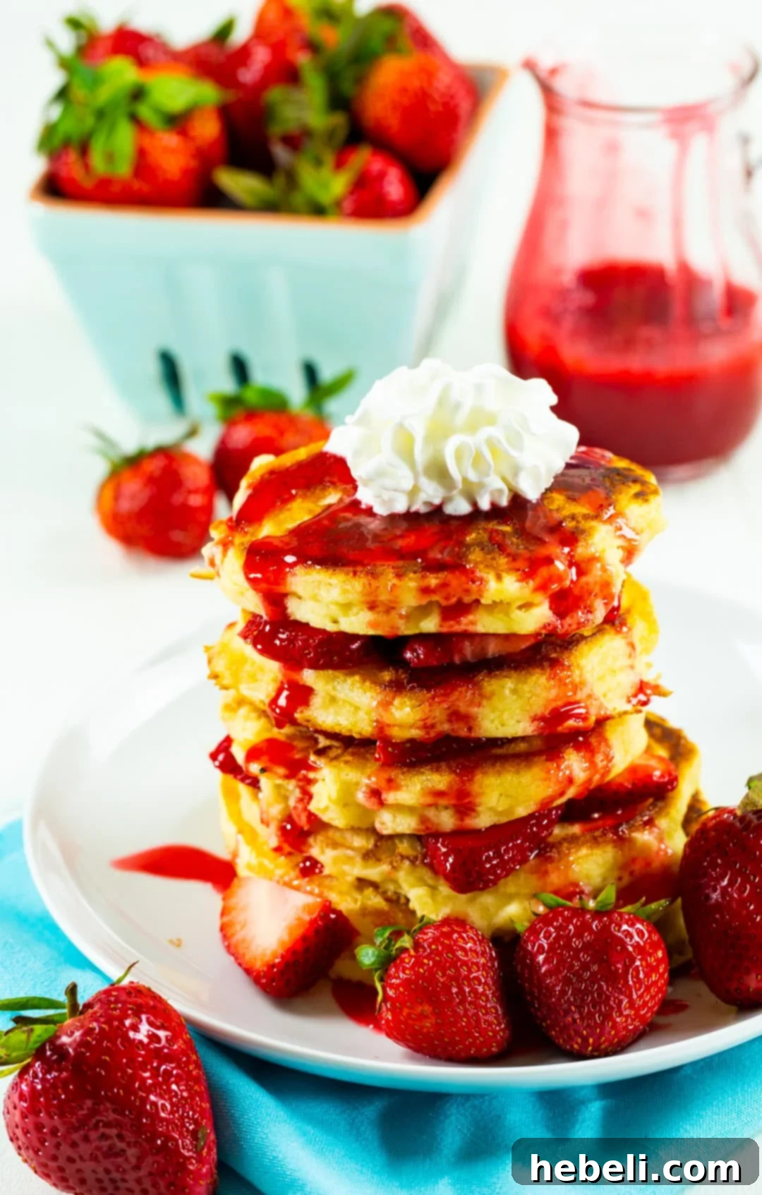 A plate of Strawberry Shortcake Pancakes adorned with whipped cream and sliced strawberries, with fresh strawberries scattered in the background.