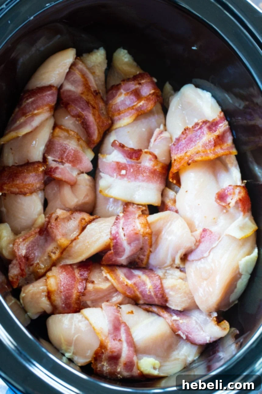 Hands wrapping partially cooked bacon around raw chicken breasts for the slow cooker.