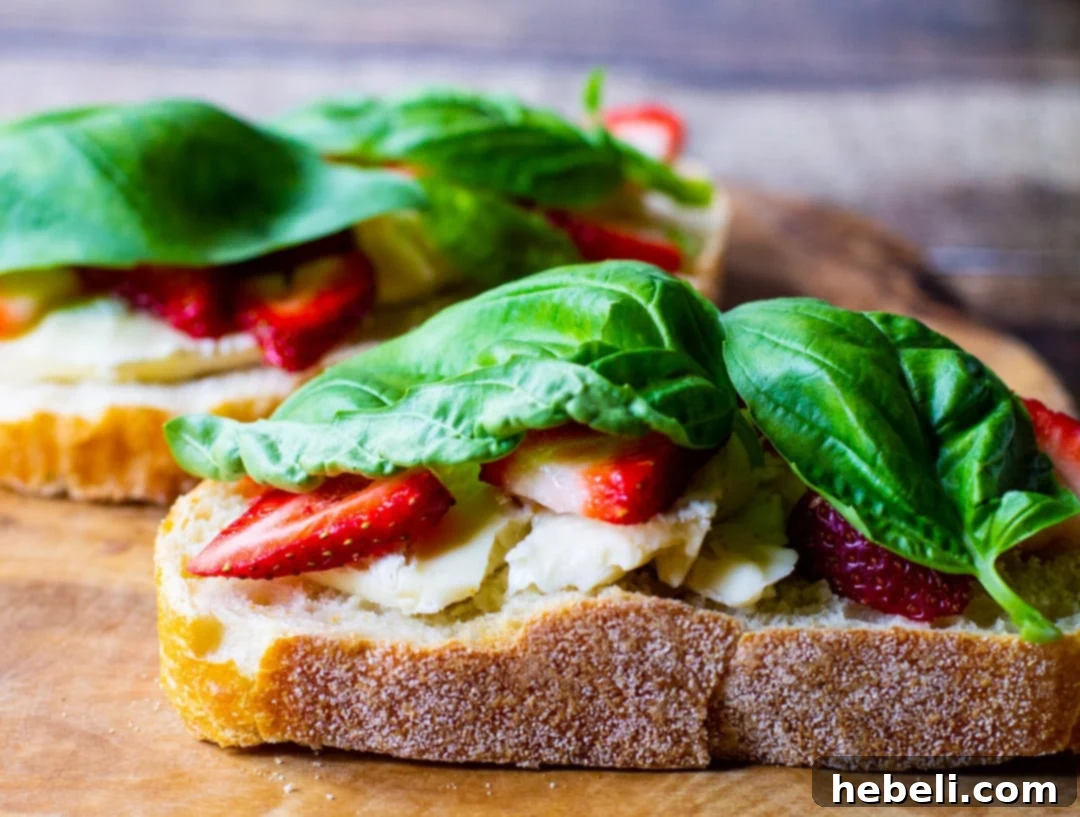 Sandwich being assembled with layers of bread, jelly, brie, strawberries, basil, and turkey.