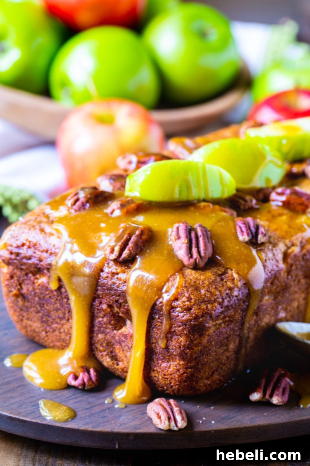 A close-up shot of a golden-brown loaf of Caramel Apple Bread, with a rustic bowl of fresh apples in soft focus behind it, highlighting the natural ingredients.