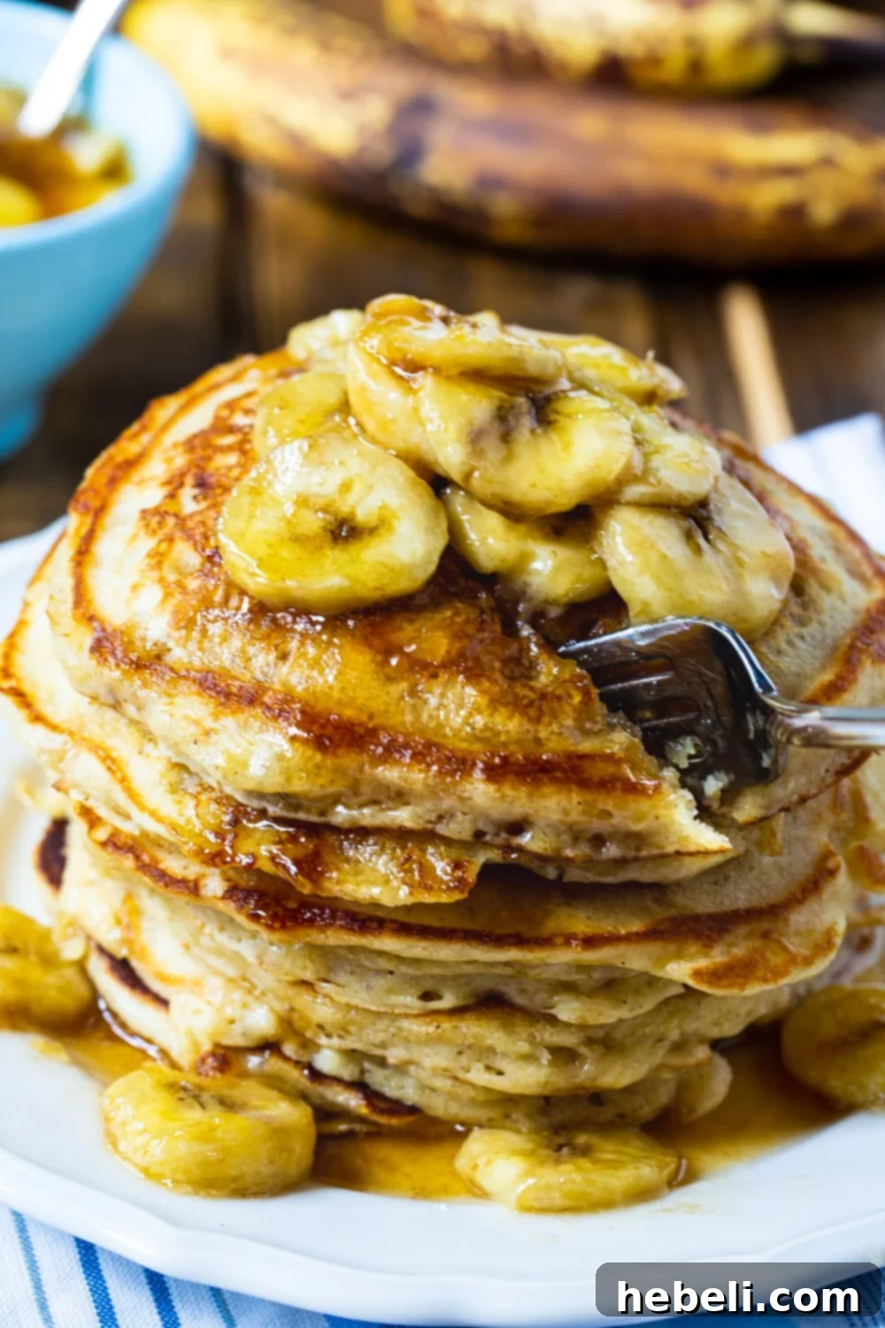 A fork cuts into a tall stack of fluffy banana pancakes, revealing their soft texture, with caramel-banana syrup dripping down the sides.