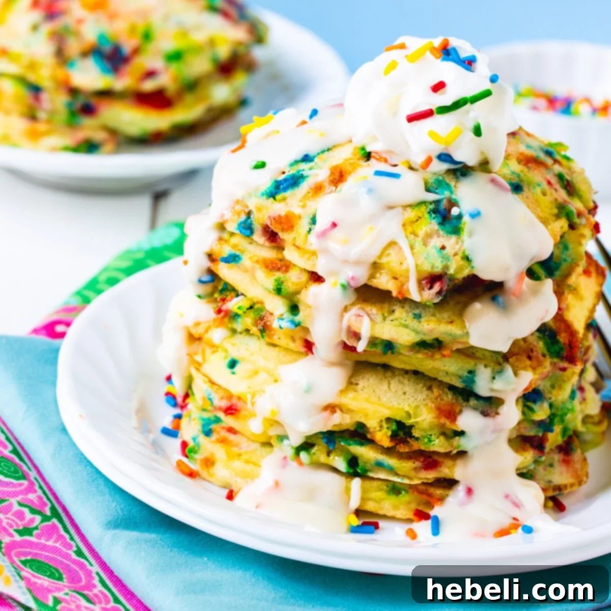 Tall stack of Cake Batter Pancakes with sprinkles and a creamy white glaze dripping down the sides, resting on a white plate with a fork.