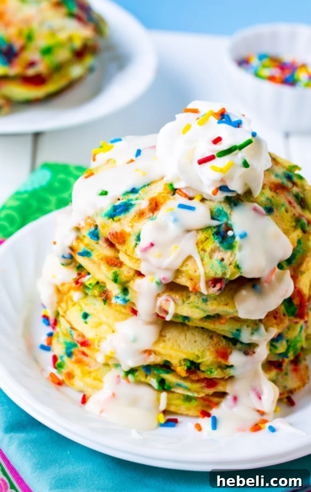 A close-up shot of a stack of fluffy Cake Batter Pancakes, generously topped with a rich, white glaze and colorful sprinkles, on a ceramic plate.