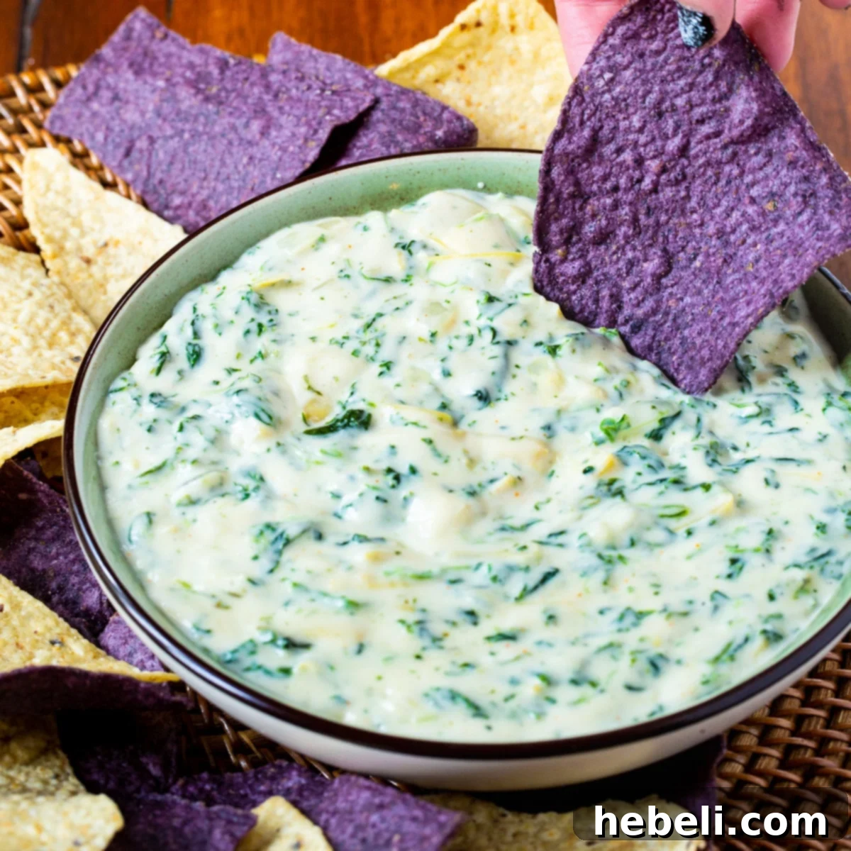 Irresistible Spinach Artichoke Dip 2 A large bowl of creamy Spinach Artichoke Dip, garnished with fresh herbs, surrounded by golden tortilla chips and toasted pita bread wedges on a wooden surface.