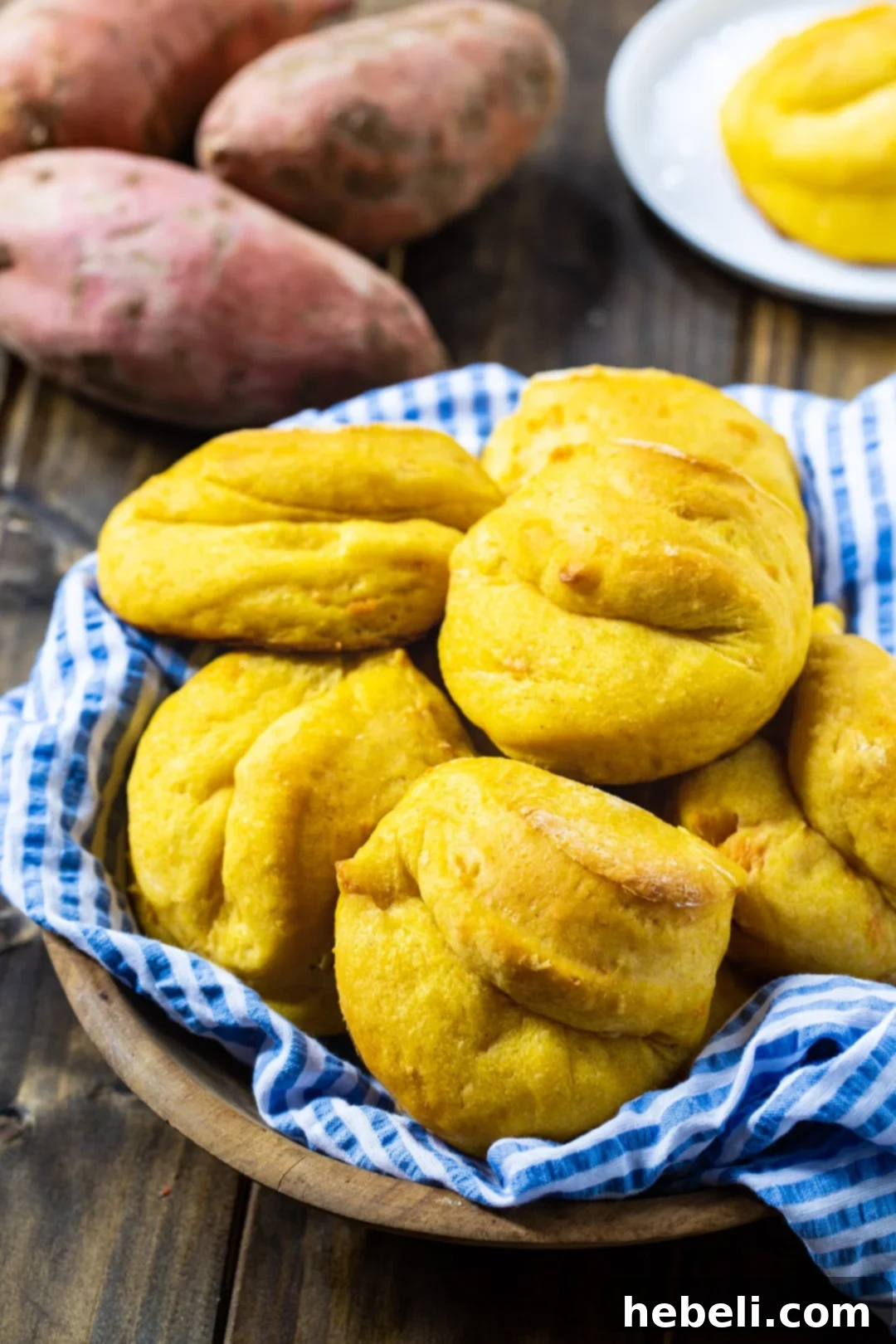 A basket overflowing with freshly baked Sweet Potato Rolls, with whole raw sweet potatoes subtly blurred in the background, hinting at their fresh ingredients.