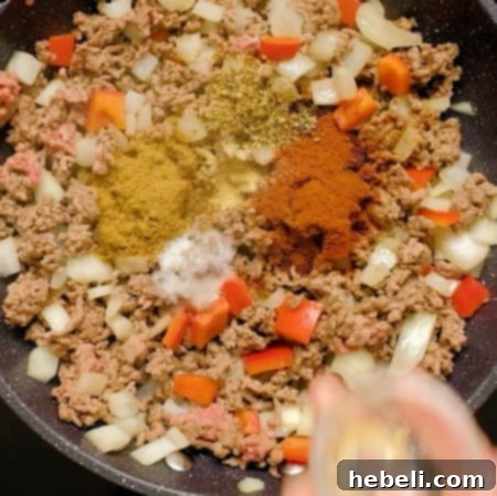 A hand adding various spices to the browned ground beef and vegetables.