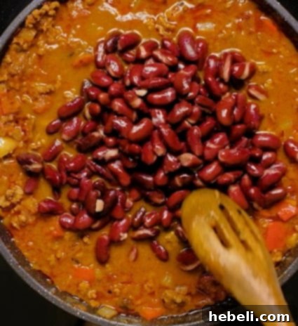 A spoon stirring kidney beans into the simmering Dr Pepper Chili.