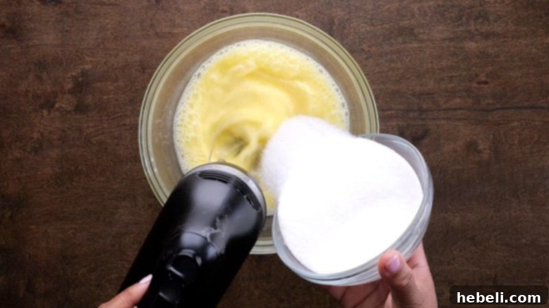 Adding granulated sugar to fluffy, beaten eggs in a mixing bowl, illustrating the aeration process.