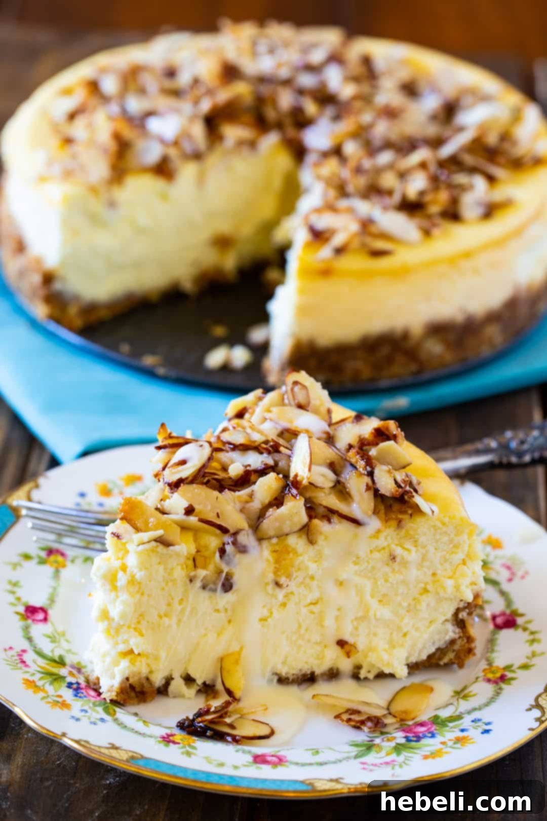 A delicious slice of Amaretto Cheesecake in the foreground, with the full cheesecake visible in the blurred background, emphasizing its creamy texture.