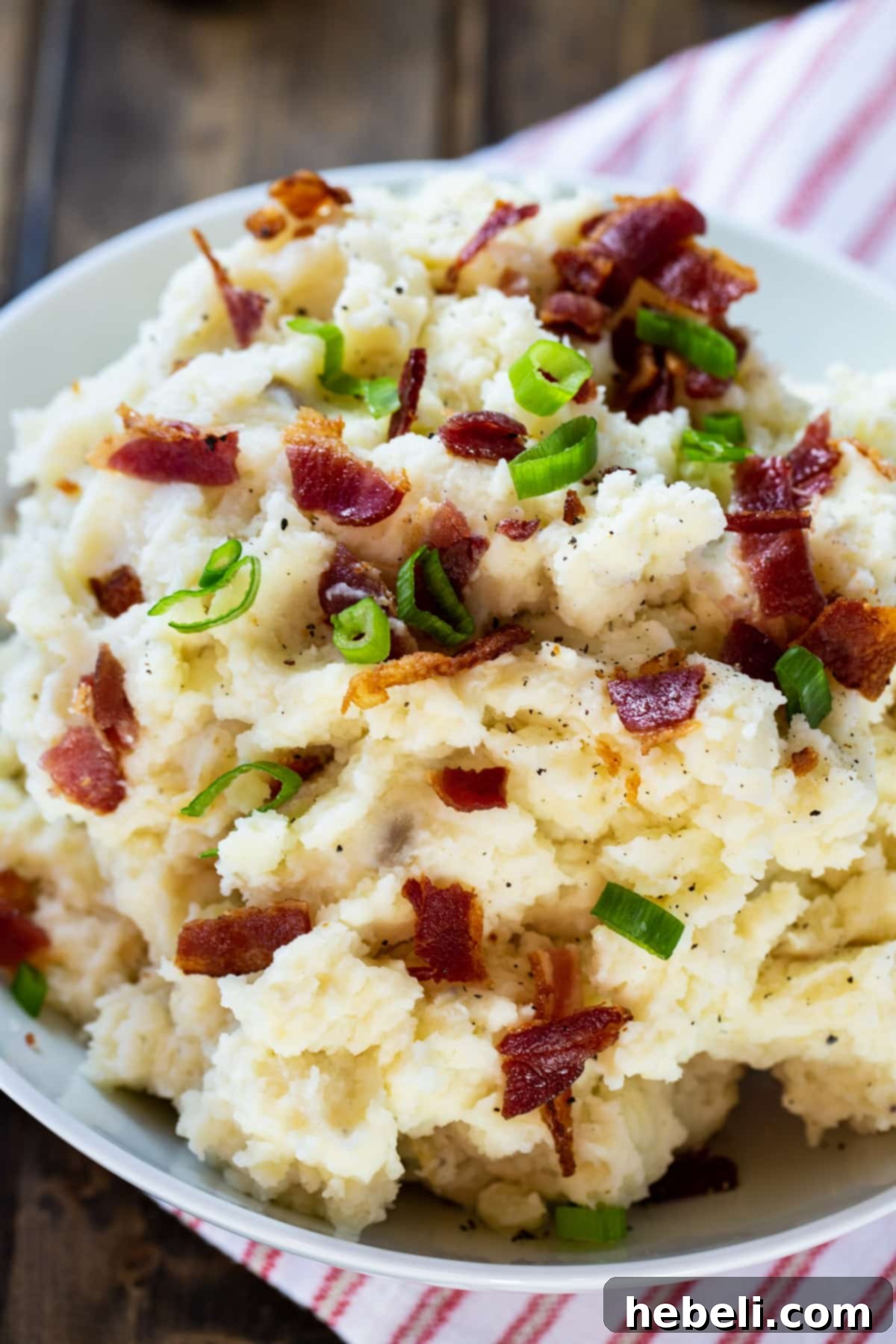 A close-up view of Roasted Garlic and Bacon Mashed Potatoes in a serving bowl, generously topped with crumbled bacon and chopped green onions.