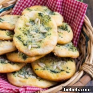 A close-up of a single Biscuit Mini Focaccia, golden brown and topped with vibrant pesto, pine nuts, and Parmesan, on a light background.