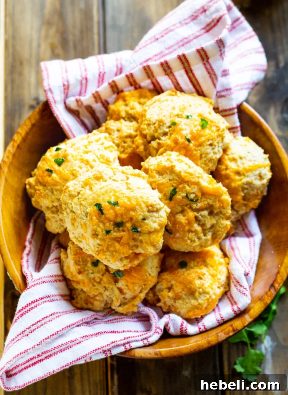 Overhead view of a rustic wooden bowl filled with golden-brown Cheddar Bay Biscuits, nestled beside a charming red and white cloth napkin.